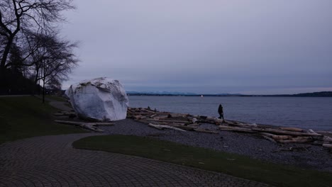 Moving-toward-a-brick-pathway-leading-to-a-giant-white-rock-at-driftwood-covered-beach-on-the-edge-of-ocean-people-walking-of-logs-on-cloudy-day-tree-grass-clouds-evening