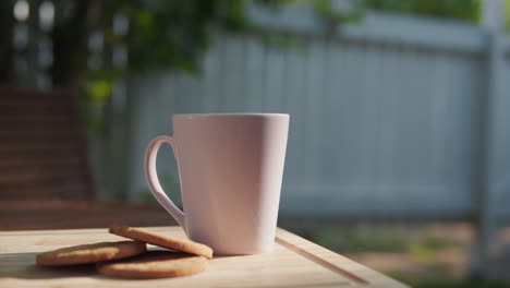 Hot-Coffee-In-White-Porcelain-Mug-With-Pieces-Of-Cookies-On-Wooden-Table-With-Sunlight
