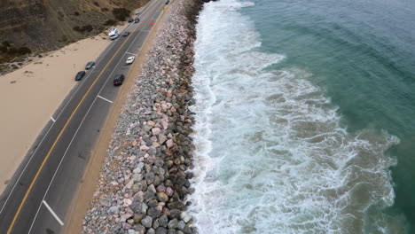 Pacific-Ocean-Waves-Crashing-on-California-Shore,-Aerial-Drone-Shot-Over-Moving-Traffic