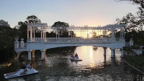 Aerial-dolly-in-of-people-in-boats-sailing-under-white-bridge-in-Rosedal-gardens-at-golden-hour,-Buenos-Aires