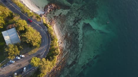 High-drone-view-looking-down-on-a-scenic-popular-swimming-area-next-to-a-coastal-road-and-car-park-area
