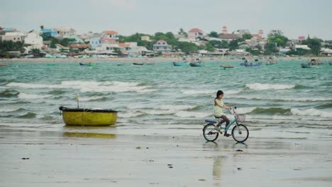 Child-girl-rides-bicycle-on-Vietnamese-sea-shore-of-Mui-Ne-beach,-near-fisherman-village