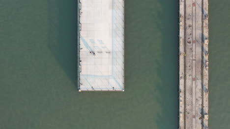 A-top-down-view-directly-above-a-concrete-pier,-next-to-an-older-pier,-taken-in-the-morning-over-a-green-river