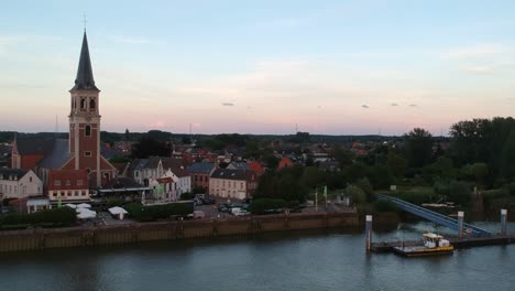 River-coastline-and-pier-with-small-town-and-church-tower,-sunset-aerial-view