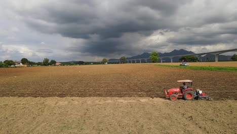 A-steady-aerial-footage-of-a-red-tractor-tilling-the-ground-during-a-cloudy-day-contrasting-the-farmland,-mountains-in-the-horizon-and-a-high-speed-elevated-railway-in-the-horizon
