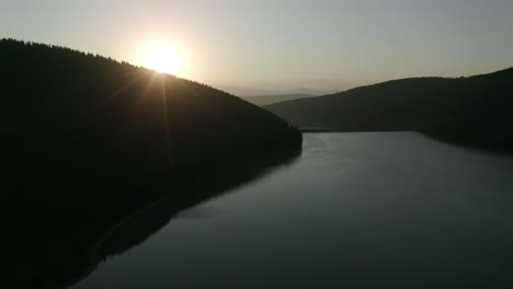 Slow-aerial-descent-towards-calm-lake,-sun-setting-behind-ridge-in-Romania