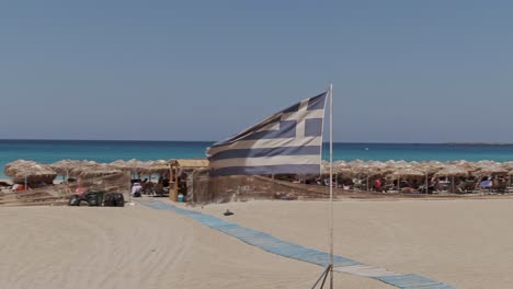 Wawing-Greece-flag-in-resort-of-Falasarna-beach-in-Crete-island,-drone-orbit-view