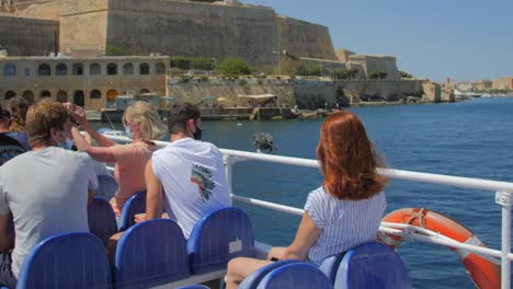Turistas-Que-Usan-Mascarilla-Mientras-Están-Sentados-En-La-Cubierta-Del-Ferry-Que-Navega-Desde-Sliema-A-Valletta,-Malta