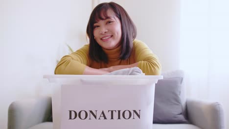 Portrait-of-young-Asian-woman-looking-at-camera-holding-box-with-clothes-for-donation