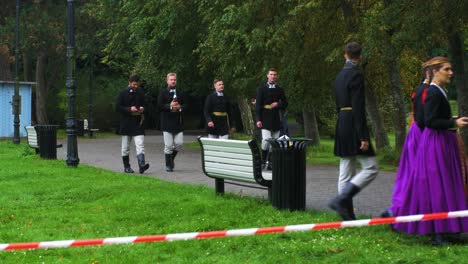 Adult-dancers-in-traditional-folk-costumes-walking-in-a-park-before-a-dance-performance-in-open-air,-sunny-summer-evening,-happy,-Latvian-national-culture,-wide-distant-shot