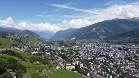 Vista-Desde-Un-Dron-De-La-Hermosa-Ciudad-De-Sion-En-Las-Montañas-Del-Sur-De-Suiza,-Hermoso-Clima-Soleado-Con-Algunas-Nubes-En-El-Cielo