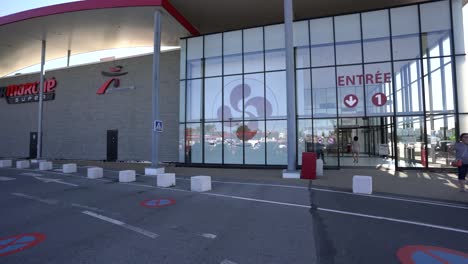 People-entering-Intermarché-Super-store-with-woman-standing-outside,-Pan-right-orbit-reveal-shot