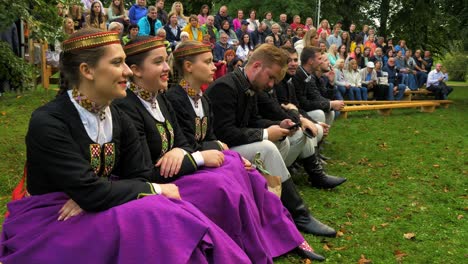 Adult-caucasian-dancers-in-traditional-folk-costumes-sitting-on-a-bench-before-a-dance-performance-in-open-air,-sunny-summer-evening,-happy,-Latvian-national-culture,-medium-handheld-shot