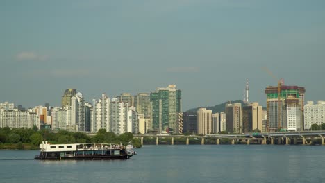 Ferry-Cruise-Ship-with-Korean-Tourists-Traveling-along-Han-River-with-Admirable-Seoul-City-Panorama---Landmark-Seoul-Namsan-Tower,-Mapodaegyo-bridge,-Gangbyeon-Expressway-Road,-Daytime-Summer