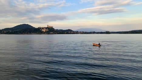 Fisherman-rowing-small-wooden-fishing-boat-on-Maggiore-lake-in-Italy-with-Angera-fortress-in-background