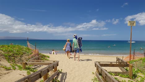 Couple-walking-onto-a-sandy-beach-carrying-a-chair,-Maui-Hawaii