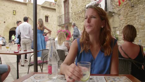 Beautiful-Young-Woman-Sitting-At-The-Cafe,-Drinking-Sparkling-Water-With-Lemon-In-Main-Square-Of-Medieval-Town-Of-Pals-In-Catalonia,-Spain