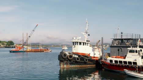 Tugboat,-trawler-and-crane-platform-anchored-along-the-coast-on-a-harbor-dock-located-in-Amador,-Panama-City-during-a-clear-sunny-summer-day-with-the-Bridge-of-the-Americas-in-the-background