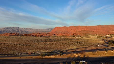 Vista-Ascendente-De-Una-Carretera-Con-Autos-Que-Pasan-En-El-Desierto-En-Un-Valle-Tranquilo-Y-Montañas-Al-Atardecer