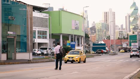 A-businessman-carrying-a-backpack-crossing-the-street-on-his-daily-commute-to-the-Fernandez-de-Cordoba-Metro-Station,-in-the-distance-the-sunsets-behind-the-beautiful-urban-cityscape,-Panama-City