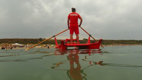 Red-Italian-lifeguard-ready-for-first-aid-emergency-standing-and-rowing-rescue-boat-while-watching-people-swimming-and-beach-in-background