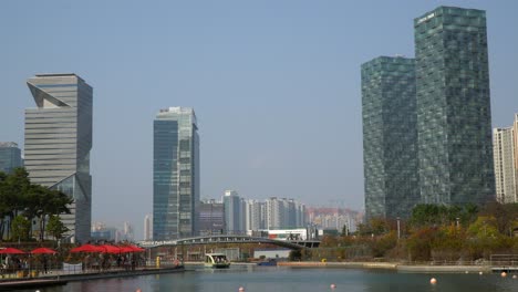 Horizonte-Urbano-Moderno-Del-Parque-Central-De-Songdo,-Torre-G---La-Gente-Viaja-En-Un-Taxi-Acuático-En-Un-Lago,-Los-Viajeros-Cruzan-Un-Puente-Peatonal