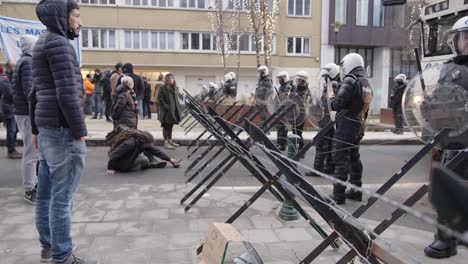 Protester-sitting-on-the-ground-facing-barbed-wire-fence-with-riot-police-officers-holding-the-line-during-anti-mandate-rally-in-Brussels,-Belgium