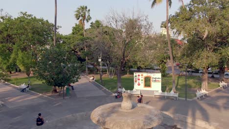 Drone-Descends-At-The-Central-Fountain-Of-Jardín-Núñez-With-People-Sightseeing-In-Colima,-Mexico