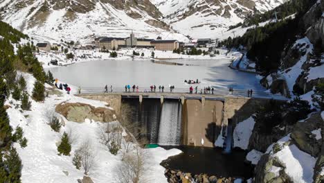 Aerial:-dam-in-a-frozen-lake-and-people-walking-along,-among-mountains-and-a-sharine-in-the-background