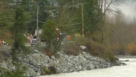 A-distant-shot-of-a-construction-team-repairing-a-damaged-road-and-river-bank-caused-by-the-devastating-floods-in-Abbotsford,-as-dump-truck-offloads-stones-to-an-excavator-in-British-Columbia,-Canada