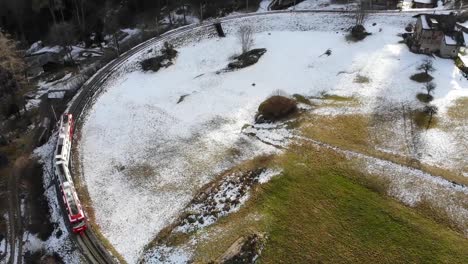 Aerial:-red-and-white-train-in-the-Alps,-passing-through-a-curve-and-some-snow