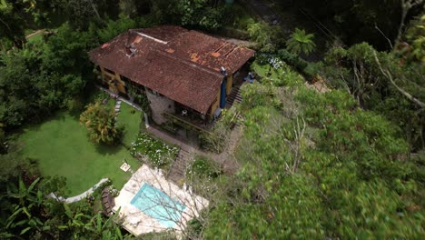 Backwards-aerial-movement-obscuring-lush-tropical-garden-greenery-with-swimming-pool-on-a-mountain-side-with-picturesque-stone-hotel-building-by-trees-in-the-foreground