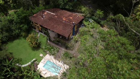 Movimiento-Aéreo-Hacia-Atrás-Que-Oscurece-El-Exuberante-Jardín-Tropical-Con-Piscina-En-La-Ladera-De-Una-Montaña-Con-Un-Pintoresco-Edificio-De-Piedra-Junto-A-Los-árboles-En-Primer-Plano