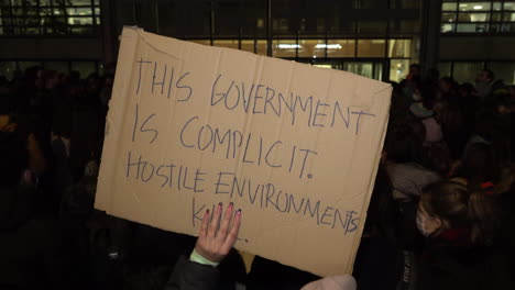 A-person-on-a-refugee-rights-protest-outside-the-UK-Home-Office-at-night-holds-up-a-cardboard-placard-that-says,-“The-government-is-complicit