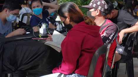 Close-up-of-Philippine-Woman-with-face-mask-reading-book-in-shopping-center-and-charging-smartphone-after-losing-home-and-electricity-cut-in-own-house