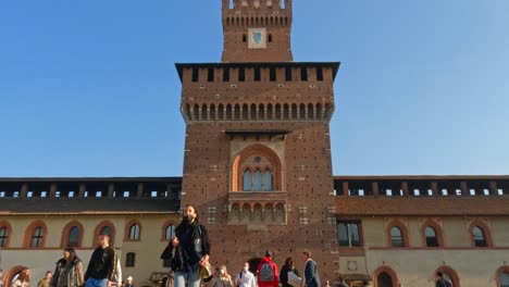 Low-angle-view-of-tourists-walking-at-Sforza-Castle-in-Milan-with-clock-tower-in-background,-Italy