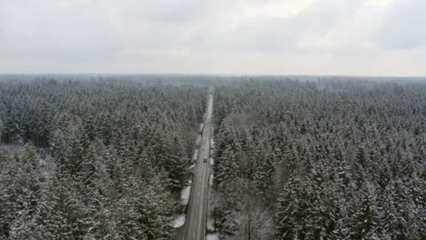 Winter-aerial-with-a-straight-street-leading-to-the-horizon,-flying-backward-to-unveil-the-wide,-big-snow-covered-treetops-of-a-conifer-forest