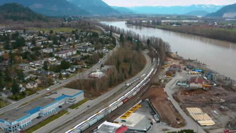 Aerial-over-Mission,-BC,-as-a-train-travels-along-the-shore-of-the-Fraser-River