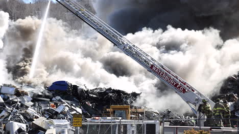 Firefighters-Use-an-Extended-Ladder-to-Spray-Water-onto-a-Burning-Scrap-Pile-with-a-Thick,-Dark-Cloud-of-Smoke-Rising-in-the-Background-on-a-Sunny-Day