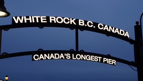 A-front-facing-view-of-the-sign-of-White-Rock-BC-Canada-World’s-longest-pier-at-night-with-people-walking-around-in-the-dark-evening-sun-setting-far-in-the-background