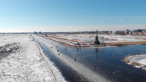 Locals-ice-skating-in-Netherlands-winter-frozen-canals,-windmill-aerial-view