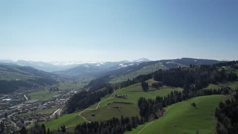 Scenic-aerial-forward-over-green-hills-in-Switzerland,-with-city-and-mountains