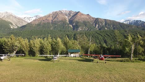 Imágenes-Aéreas-De-Camiones-Disparados-A-Lo-Largo-De-Aviones-Estacionados-A-Lo-Largo-De-Una-Pista-De-Aterrizaje-De-Hierba-En-La-Cordillera-Talkeetna-De-Alaska