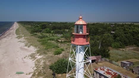 Beautiful-aerial-view-of-white-painted-steel-lighthouse-with-red-top-located-in-Pape,-Latvia-at-Baltic-sea-coastline-in-sunny-summer-day,-wide-angle-ascending-drone-shot-moving-forward-close