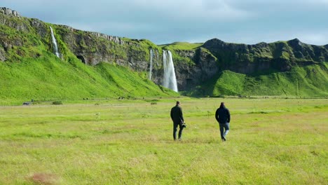 Two-Tourists-walking-towards-Seljalandsfoss-and-Gljufrabui-waterfalls-over-a-beautiful-field-of-grass-on-a-sunny-day-in-Southern-Iceland