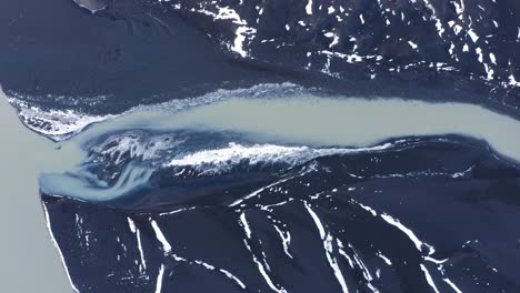 Luftaufnahme-Von-Oben-Nach-Unten,-Wie-Ein-Gletscherfluss-Ins-Meer-Fließt,-Umgeben-Von-Vulkanlandschaft-Und-Schnee-Im-Winter-In-Island