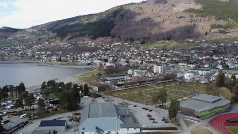 Vossevangen-town-centre-with-Vossabadet,-Vangsvatnet-lake,-sports-arena-and-graveyard---Spring-at-Voss-Norway---Reverse-aerial