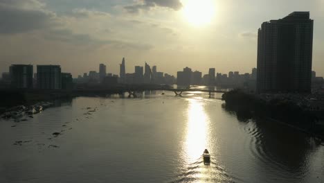tracking-a-traditional-river-boat-along-the-Saigon-river,-Vietnam-from-drone-with-view-to-modern-city-skyline-at-sunset