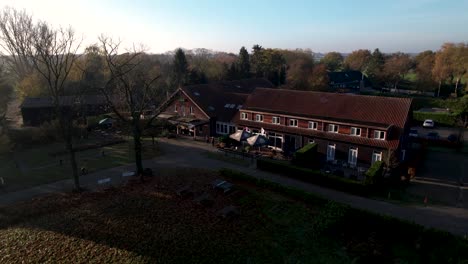 Luftaufnahme-Der-Niederländischen-Herbstlandschaft-Bei-Sonnenaufgang-Am-Frühen-Morgen-Mit-Langen-Schatten-Von-Bäumen-Rund-Um-Das-Hotelgebäude-In-Der-Landschaft-Der-Niederlande