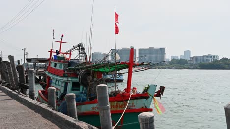 A-boat-with-a-red-flag-docked-and-Pattaya-City-is-at-the-background,-Pattaya-Fishing-Dock,-Chonburi,-Thailand